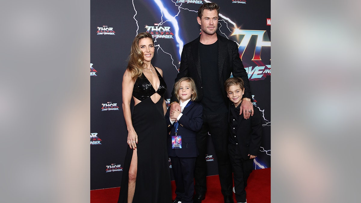 Chris Hemsworth con camisa y chaqueta negras sonríe con sus dos hijos (uno con un traje negro completo, el otro con un traje negro y camisa blanca) y su esposa Elsa Pataky con un vestido de corte negro en la alfombra roja.