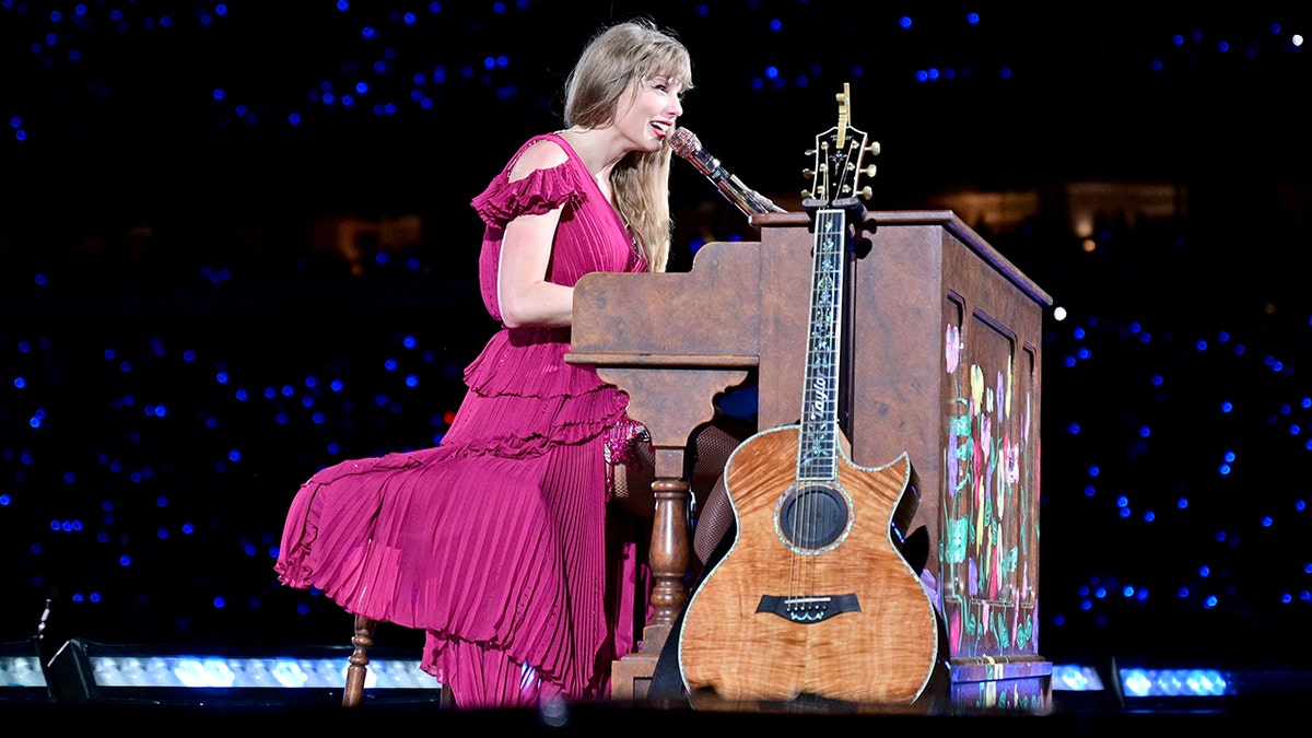 Taylor Swift con un vestido rosa actúa detrás de un piano marrón con una guitarra inclinada en el Eras Tour.