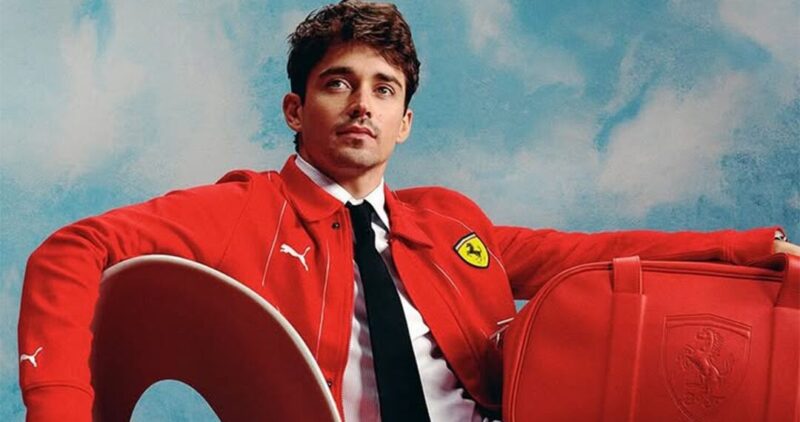 Un hombre vestido con una chaqueta roja de Ferrari está sentado en una silla junto a un bolso rojo de Ferrari, lo que ejemplifica el lujo.