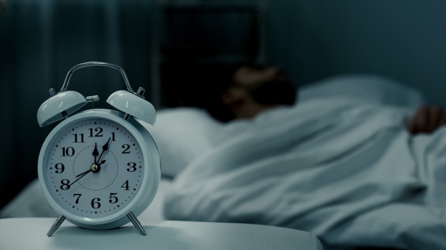 Un despertador frente a una persona tenue en la cama.