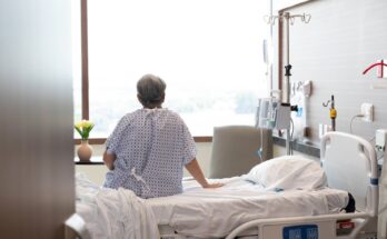 An elderly woman sits on a hospital bed with an IV drip