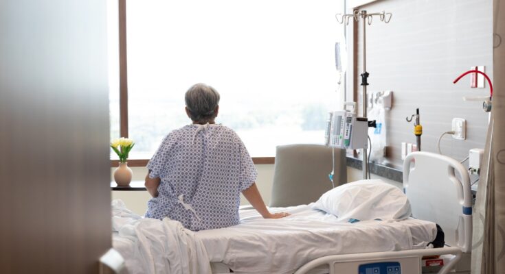 An elderly woman sits on a hospital bed with an IV drip