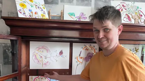 Alex se paró cerca de su obra de arte que es un puntero rosado a la parrilla rosada con comparaciones extraídas. Tienes cabello gris azul y una camiseta amarilla.