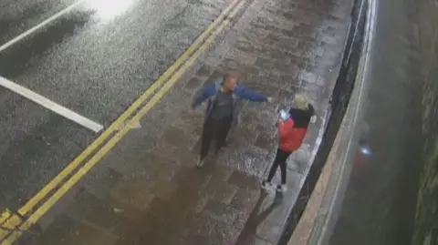 Michael CccToage oposición a cocinar con Carol O'Reilly a la calle. Señala su cara, con una chaqueta roja y negra y con una chaqueta azul 
