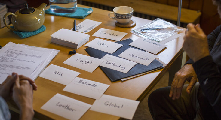 Flashcards spread out across a table as two people sit on either side of it, one with hands clasped, the other with hands resting naturally in front of them. Only their hands are visible and not the rest of their bodies.