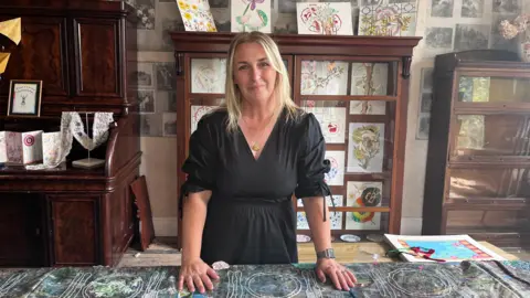 Beth tiene el cabello molila largo, con un vestido negro y sonriendo en la cámara. Rodeado de antigüedades y antiguas y antiguas de tabla de madera con imágenes. 