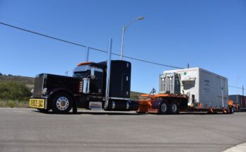 A long, dark purple semi-truck with chrome accents and a yellow "OVERSIZE LOAD" banner on the front bumper is parked on a paved road. The truck is hauling a large piece of cargo on a trailer, which is covered by a white tarp with the "AIRBUS" logo. Several tall, light-green gas cylinders are strapped to the front of the cargo. The scene takes place on a clear, sunny day with rolling, grassy hills in the background.