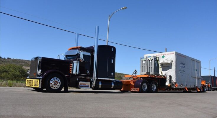 A long, dark purple semi-truck with chrome accents and a yellow "OVERSIZE LOAD" banner on the front bumper is parked on a paved road. The truck is hauling a large piece of cargo on a trailer, which is covered by a white tarp with the "AIRBUS" logo. Several tall, light-green gas cylinders are strapped to the front of the cargo. The scene takes place on a clear, sunny day with rolling, grassy hills in the background.