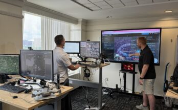 Two men stand in an office looking at four smaller computer screens and one larger computer screen. There is a tan desk in the foreground housing two computer monitors and a small grey drone. In the background stands a man in a white polo shirt facing two other computer monitors on a desk. Behind this set up is a large TV monitor with a man standing with his arm reached out in front. The man is wearing a black polo shorts and tan shorts as well as white tennis shoes. The screen shows a map and different pieces of software working in unison.