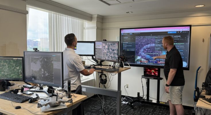 Two men stand in an office looking at four smaller computer screens and one larger computer screen. There is a tan desk in the foreground housing two computer monitors and a small grey drone. In the background stands a man in a white polo shirt facing two other computer monitors on a desk. Behind this set up is a large TV monitor with a man standing with his arm reached out in front. The man is wearing a black polo shorts and tan shorts as well as white tennis shoes. The screen shows a map and different pieces of software working in unison.