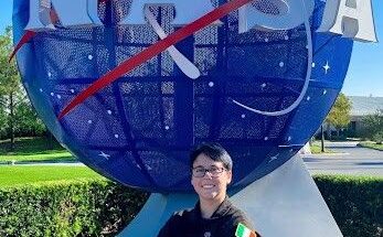A person wearing a black NASA flight suit stands smiling with arms crossed in front of the large blue NASA globe sculpture. The Irish flag patch is visible on their sleeve, and the sky is clear and blue in the background.