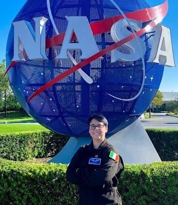 A person wearing a black NASA flight suit stands smiling with arms crossed in front of the large blue NASA globe sculpture. The Irish flag patch is visible on their sleeve, and the sky is clear and blue in the background.