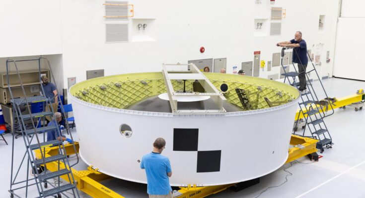 Image shows a white and yellow cylinder piece of hardware with technicians inspecting and taking photos of the Orion stage adapter at NASA's Kennedy Space Center in Florida. Photo credit: NASA/Kim Shiflett