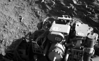 A grayscale photo from the Martian surface shows rocky, uneven terrain in medium gray. Part of the Curiosity rover fills most of the bottom half of the image, brightly lit in some places and shadowed in others. In front of it, visible in the upper right corner of the image, a virtual wall composed of stacked, jagged, horizontal layers of stone; this abutment appears to block the rover’s path.