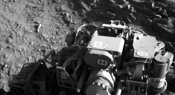 A grayscale photo from the Martian surface shows rocky, uneven terrain in medium gray. Part of the Curiosity rover fills most of the bottom half of the image, brightly lit in some places and shadowed in others. In front of it, visible in the upper right corner of the image, a virtual wall composed of stacked, jagged, horizontal layers of stone; this abutment appears to block the rover’s path.