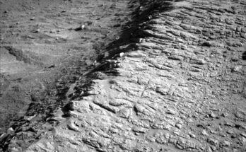 A grayscale photo from the Martian surface shows rough terrain covered in small rocks, divided across the middle of the image by a ridge running from the lower left corner of the frame to the upper right corner. The ground to the right of the ridge is relatively flat, while to the left it falls away from the ridge into a depression. The entire scene is light to medium gray, except for a very dark shadow along the left side of the ridge.