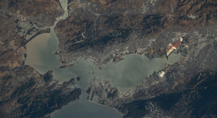 California's San Francisco Bay Area as seen from the International Space Station orbiting 260 miles above. The water is slate gray, while the surrounding land is in shades of brown, deep green, and gray.