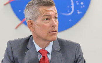 a man with gray hair and pale complexion wears a gray suit and red tie while sitting at a table under a red, white and blue NASA logo on the wall behind him