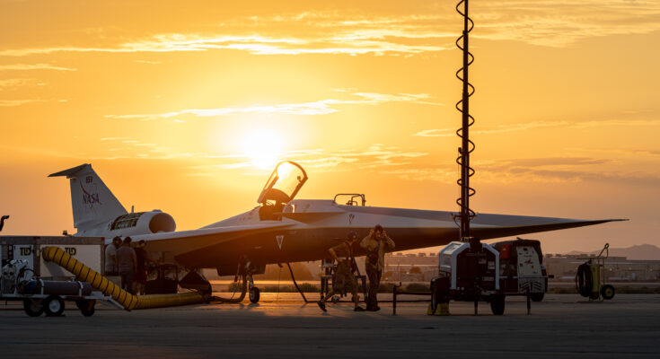 NASA’s X-59 quiet supersonic research aircraft is seen on the ramp at sunrise with its canopy open. Orange and gold light filters through the clouds behind the aircraft, highlighting the aircraft’s shape. Several ground crew members are visible around the aircraft, along with support carts. A vertical light pole with a coiled cable can be seen in the foreground.