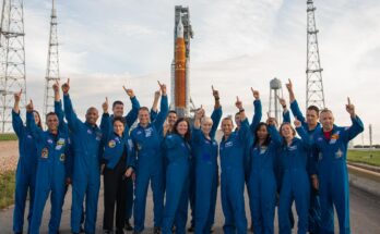 a bunch of astronauts in flight suits standing in front of a huge rocket and pointing at the sky