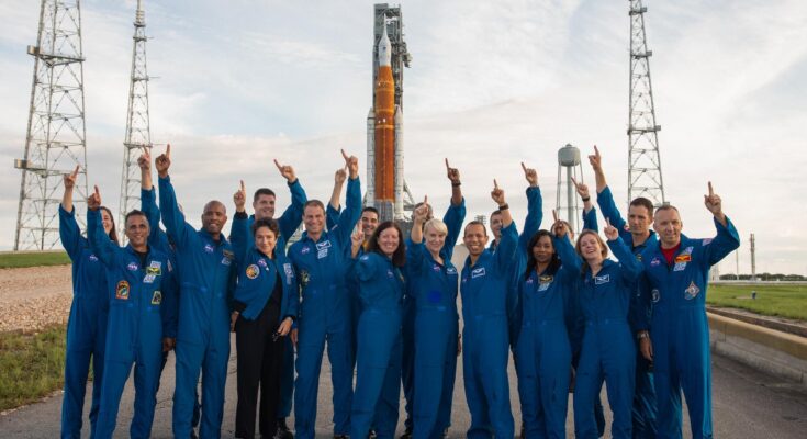 a bunch of astronauts in flight suits standing in front of a huge rocket and pointing at the sky