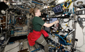 a woman in a forest green short sleeve shirt, kahki slacks and a red sweatshirt tied around her waist works with several laptop computers inside a space station module