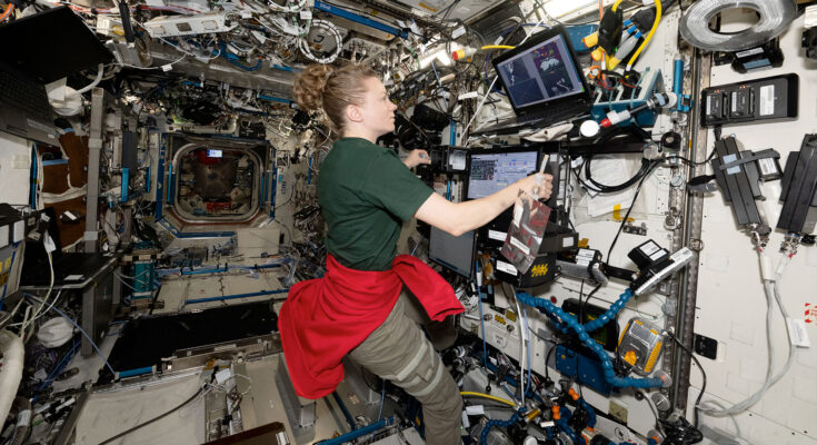 a woman in a forest green short sleeve shirt, kahki slacks and a red sweatshirt tied around her waist works with several laptop computers inside a space station module