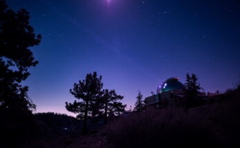 A faint beam stretches up and to the left from the open dome of a laboratory into the sky. The building and surrounding trees are mostly in shadow. The sky is a deep blue with multicolored stars visible; the bottom of the sky where it meets the landscape fades from blue to pink.