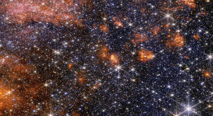 A wide view of a region of space filled with stars and clumps of orange clouds.