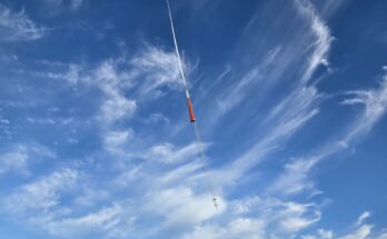 A large white scientific balloon in the shape of an upside down tear drop floats high in a bright blue sky with wispy white clouds, suspended by a long tether line connected to a white  vehicle with a crane visible on the ground below in an open field. The crane is holding the BOOP experiment.