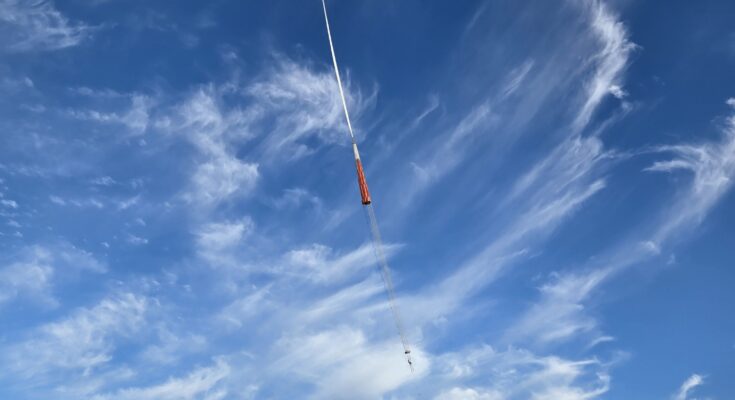 A large white scientific balloon in the shape of an upside down tear drop floats high in a bright blue sky with wispy white clouds, suspended by a long tether line connected to a white  vehicle with a crane visible on the ground below in an open field. The crane is holding the BOOP experiment.