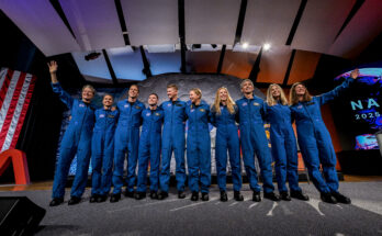 Ten people in blue jumpsuits link arms. The two women on either end wave at the audience, who is not visible.