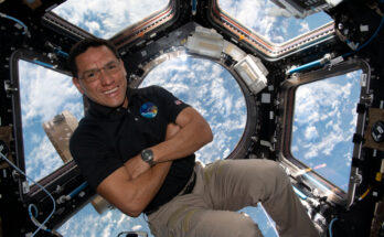 A man in a dark polo shirt smiles at the camera with his arms crossed. He is in the cupola of the International Space Station, an area with multiple windows through which Earth and space can be seen. Earth's clouds can be seen through the windows behind him.