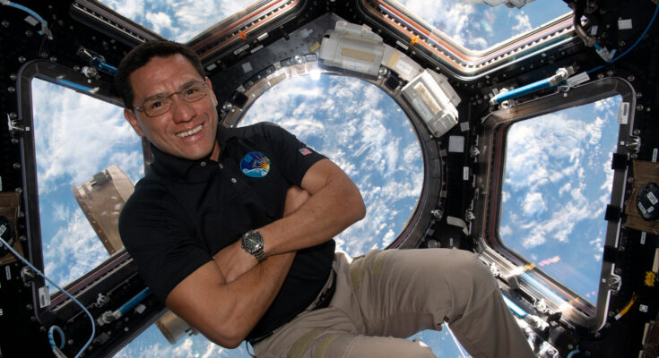 A man in a dark polo shirt smiles at the camera with his arms crossed. He is in the cupola of the International Space Station, an area with multiple windows through which Earth and space can be seen. Earth's clouds can be seen through the windows behind him.