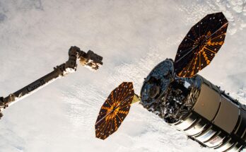 A cylinder-shaped spacecraft with prominent cymbal-shaped solar arrays on either side of it nears a long robotic arm, with a cloudy Earth below.