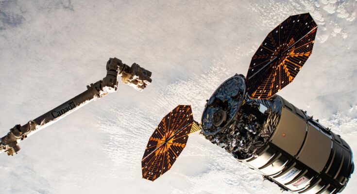 A cylinder-shaped spacecraft with prominent cymbal-shaped solar arrays on either side of it nears a long robotic arm, with a cloudy Earth below.