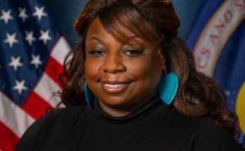 A woman poses in front of a blue background with the U.S. flag (left) and NASA flag (right) behind her.