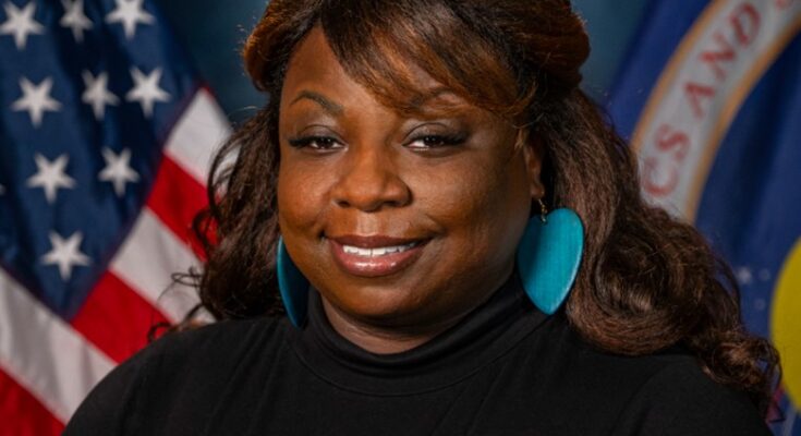 A woman poses in front of a blue background with the U.S. flag (left) and NASA flag (right) behind her.