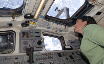 A woman wearing a headset and a green long sleeve shirt looks out of the windows on the International Space Station aimed at Earth