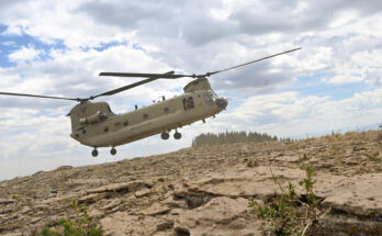 A helicopter with dual propellors floats over the dry, weedy Colorado landscape with a large cloudy sky above.