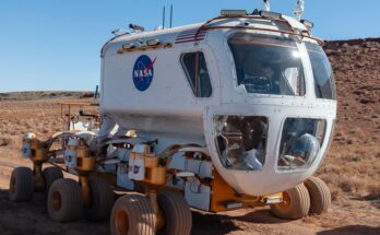 A white vehicle with six wheels and a rounded front window rolls across a dusty surface.