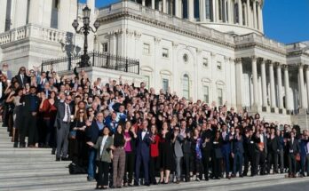 A large group of people stand on the steps of the U.S. Capitol in bright sunlight.