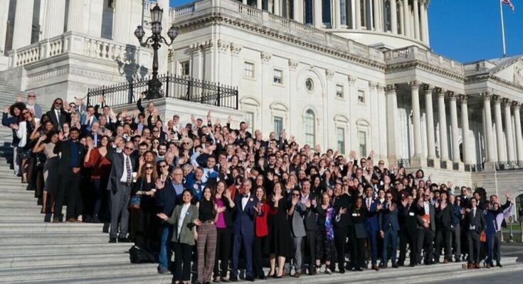 A large group of people stand on the steps of the U.S. Capitol in bright sunlight.