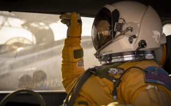 A person wears a tan astronaut suit and helmet and sits in the passenger seat of a car while a plane sits behind them on a tarmac.