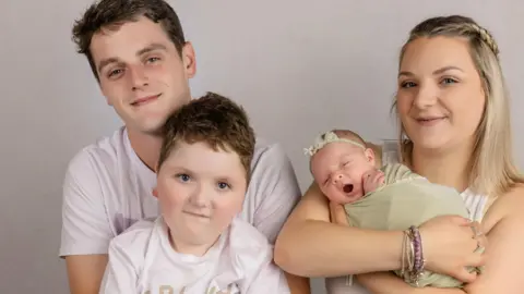 Al fotografiar a Nino Traval, su hijo de cinco años y su hija pequeña pusieron el apellido frente a un fondo gris. Mi padre, a la izquierda, tiene el pelo rubio y una camiseta blanca. Su hijo se sienta frente a él y tiene el pelo azul claro y una camiseta a juego. Yo y yo tenemos cabello largo, hermoso, blanco, dorado y morado. Él sostiene a una niña que lleva una diadema verde y una diadema verde.