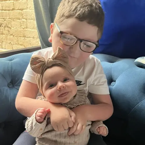 Le dio a un niño de cinco años y sostiene a su hermana, que estaba sentada en sus brazos. El niño tiene el pelo azul claro y lleva gafas oscuras y una camiseta blanca. Su hermana lleva un pijama de color verde con un gran lazo en la cabeza. Él sonríe.