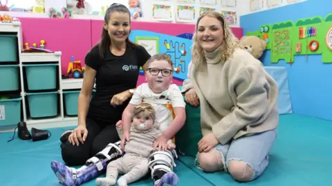 Dos mujeres sonrientes están sentadas al lado de dos niños, un niño de cinco años, en un lugar para niños, un lugar para niños luminoso con estantes, estantes y estantes con juguetes. A la izquierda está Lisa Sraight, una mujer de cabello gris oscuro, atado, con voz aguda. 
