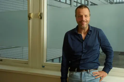 Presenta a un hombre de cabello castaño y la luz del día es cercana mientras se encuentra frente a dos grandes ventanas con marcos coloridos. Viste una camisa azul claro y pantalones azul claro y apoya su mano izquierda en su regazo izquierdo.