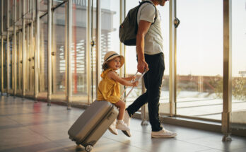 Cheerful husband and his anonymous wife walking with their little girl sitting on luggage at the airport.