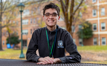 Daniel Meng-Saccoccio sits outside at a table smiling at the camera.